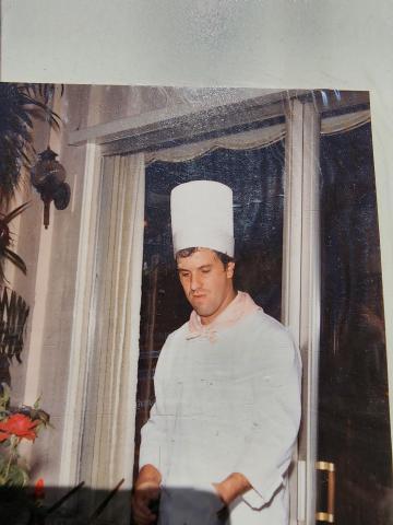 Photo of Chef Skinner works an outdoor event in full chef's whites uniform, including a tall toque blanche and pink bandana.