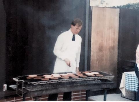 Blurry photo of David Edwards in an apron cooking burgers on a long grill at an Ath event.