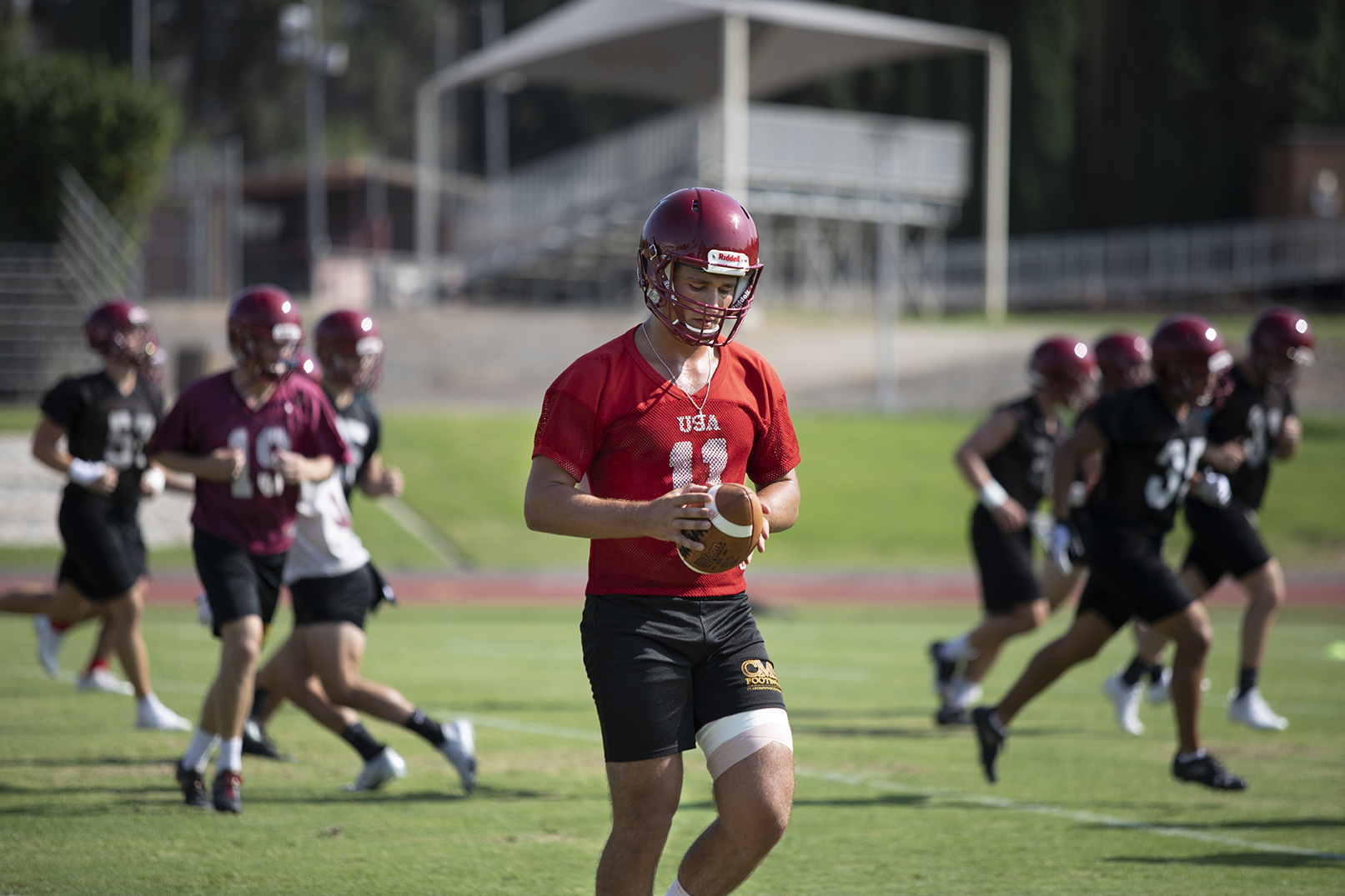 Hank Harvego '23, geared in helmet and practice jersey, hold a football while teammates run drills behind hi on John Zinda Field.