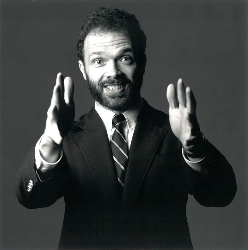 Black and white portrait of Professor Jack Pitney smiling with his arms enthusiastically raise toward camera