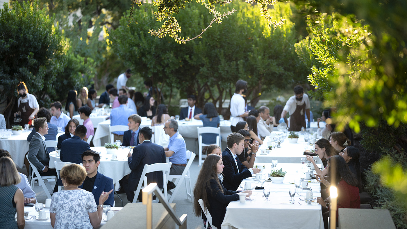 Ath attendees smile, eat, and engage with each other at their outdoor dinner tables as the sun sets over the surrounding trees.