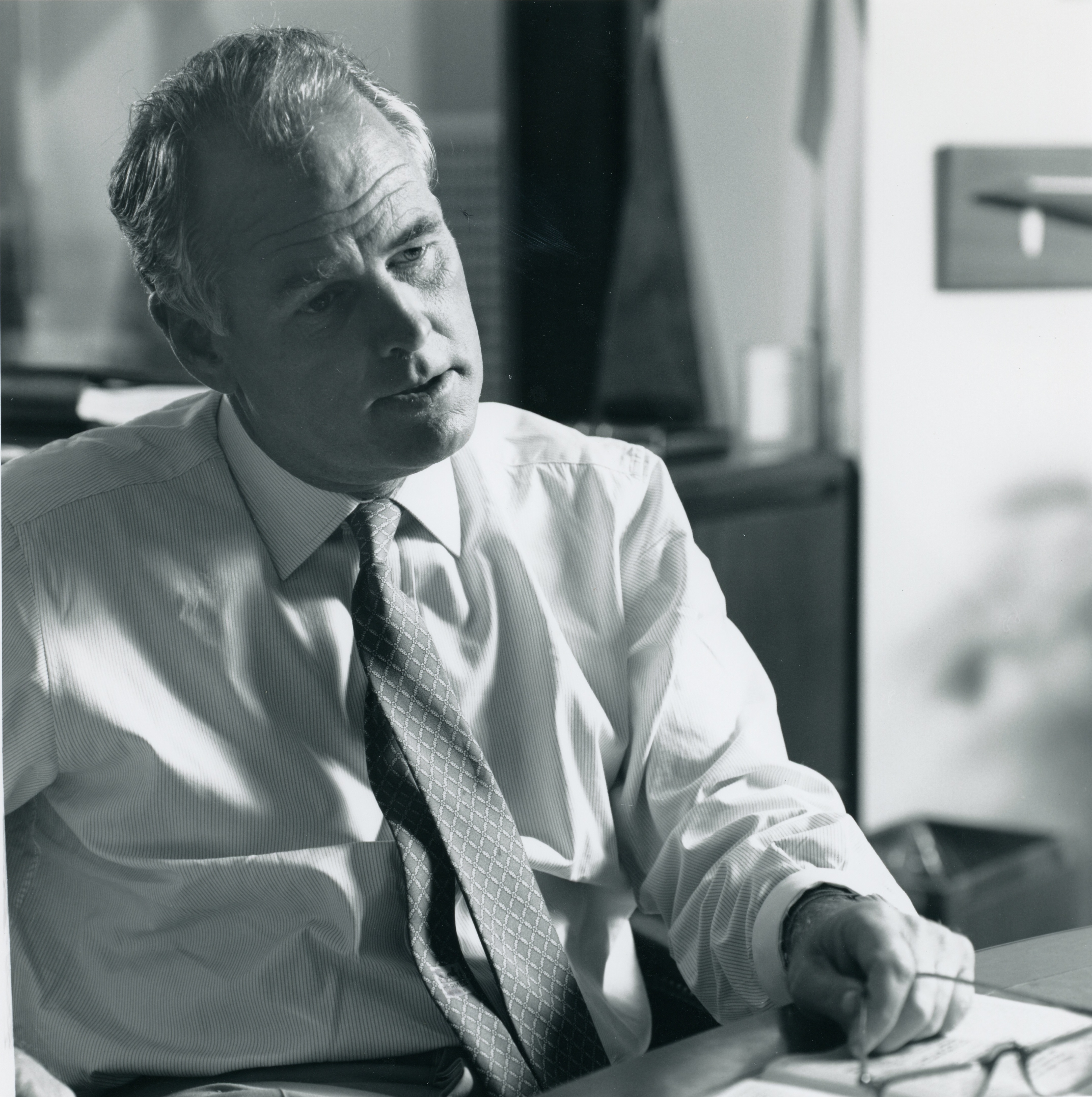 Black and white candid photo of Robert Day in March 1990. He wears a white dress shirt, a tie, and is holding his glasses by the legs while he addresses someone off camera.