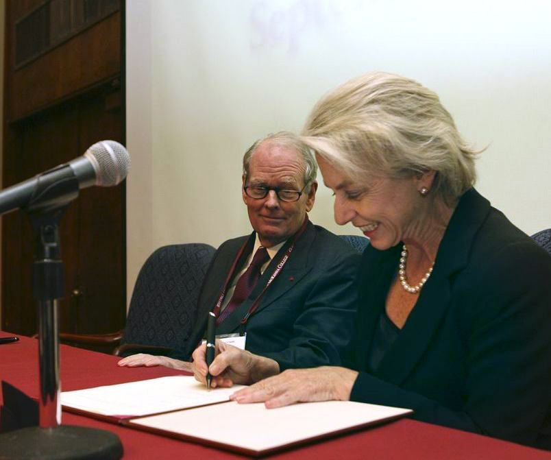 On a red conference table equipped with a mic, then CMC President Pamela Gann in a suit and pearl necklace signs off on Robert's historic gift to the school while he looks on seated beside her.