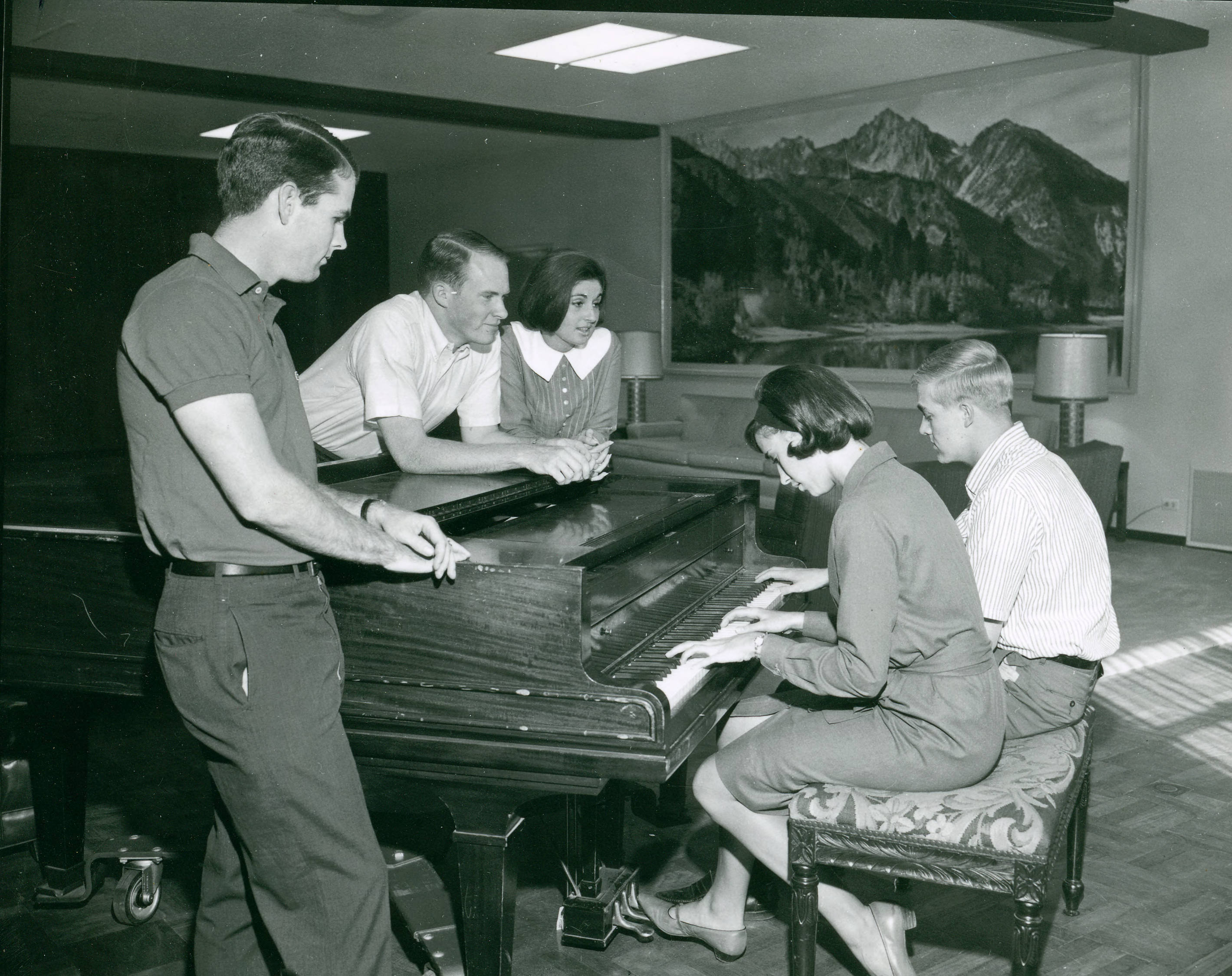Black and white photo of Robert Day and friends gathered around a piano where two people are seated and playing.