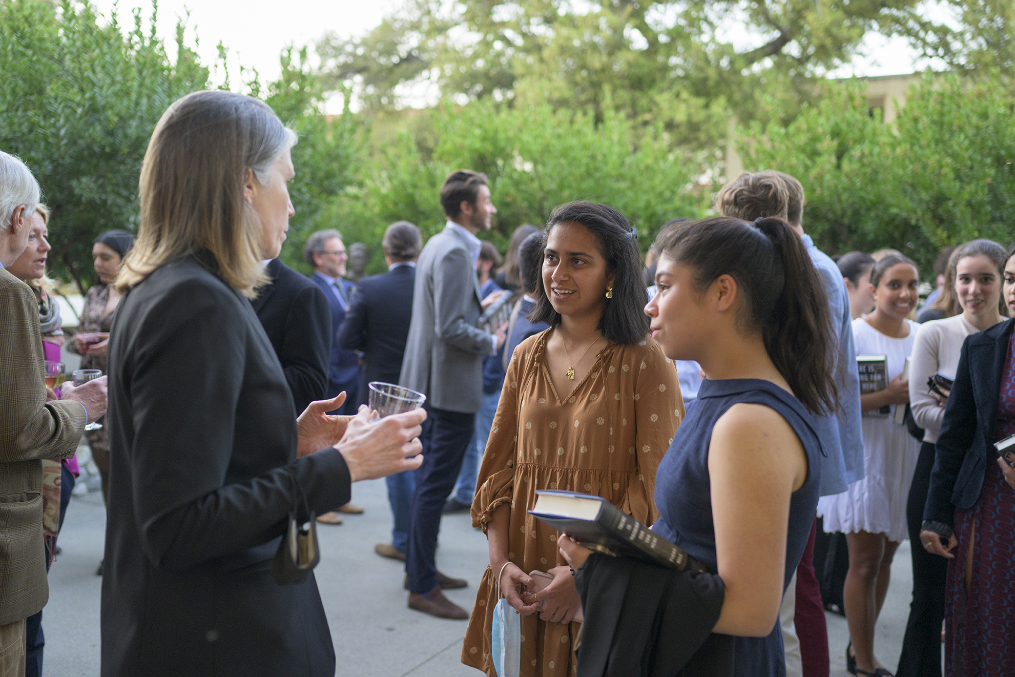 Fiona Hill speaks to two students on the Ath patio. Behind them, a line of students hold copies of her book and faculty mingle with other attendees.
