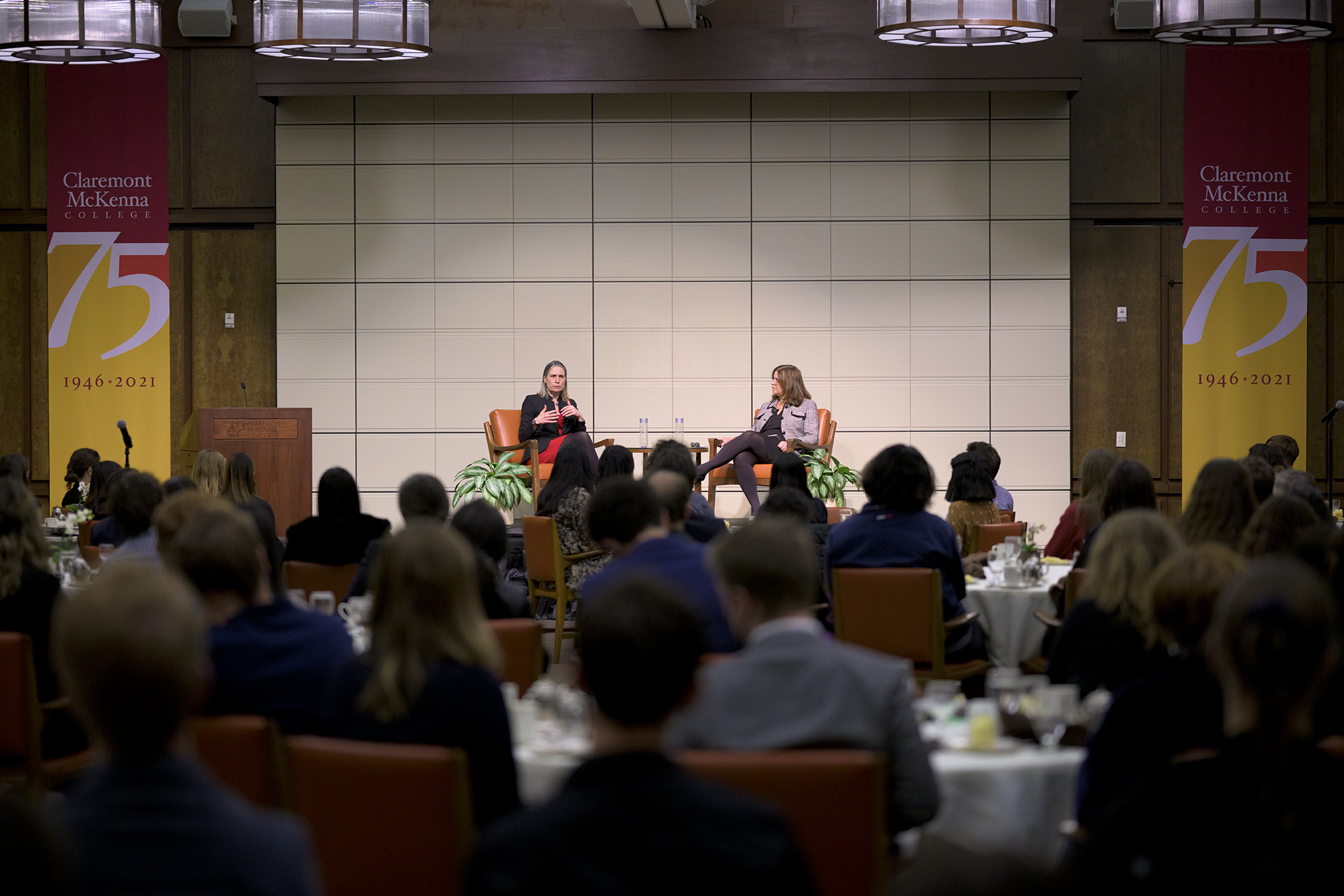 In the fore, rings of students are seated inside the Ath looking ahead at the stage. In the back, Dr. Fiona Hill and Prof. Hilary Appel speak on stage.