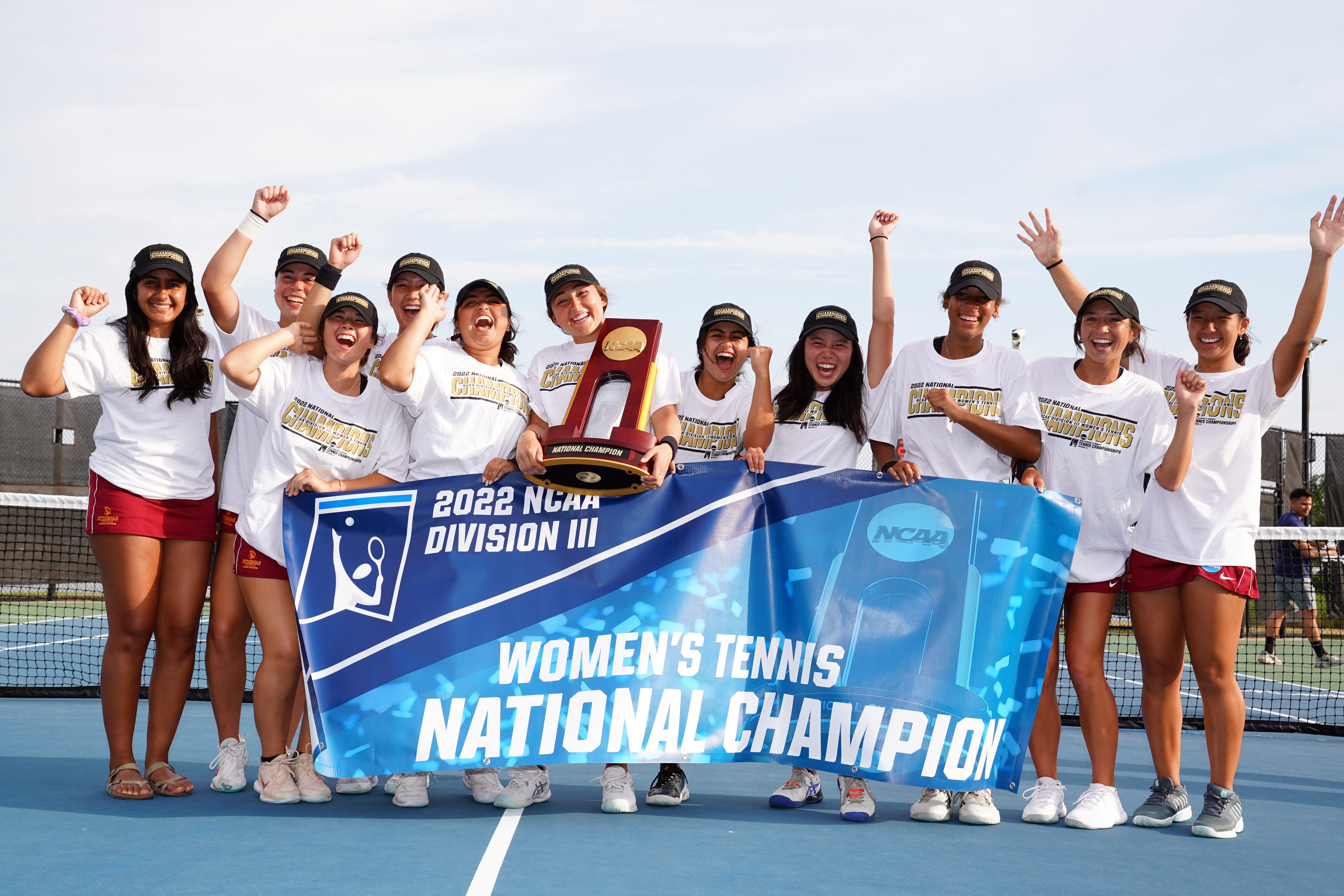 Athena tennis team, geared up in championship t-shirts and baseball caps, holds up the 2022 NCAA Division III Women's Tennis National Championship banner and award trophy.