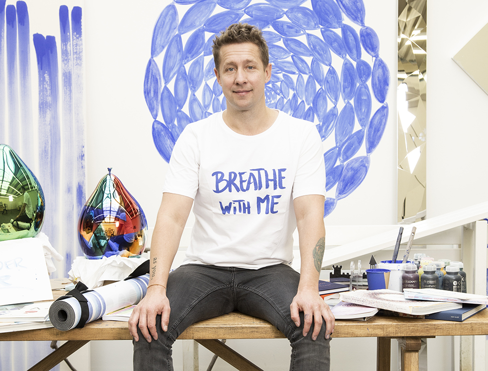 Artist Jeppe Hein sitting on a wooden table surrounded by his mirror balloon pieces and bottles of paints and brushes. Behind him is a partial view of his collaborative Breathe With Me project.