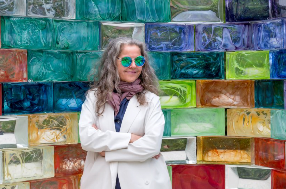 Portrait of the artist Pae White standing before a wall of rainbow colored glass bricks, all part of her piece Qwalaa.