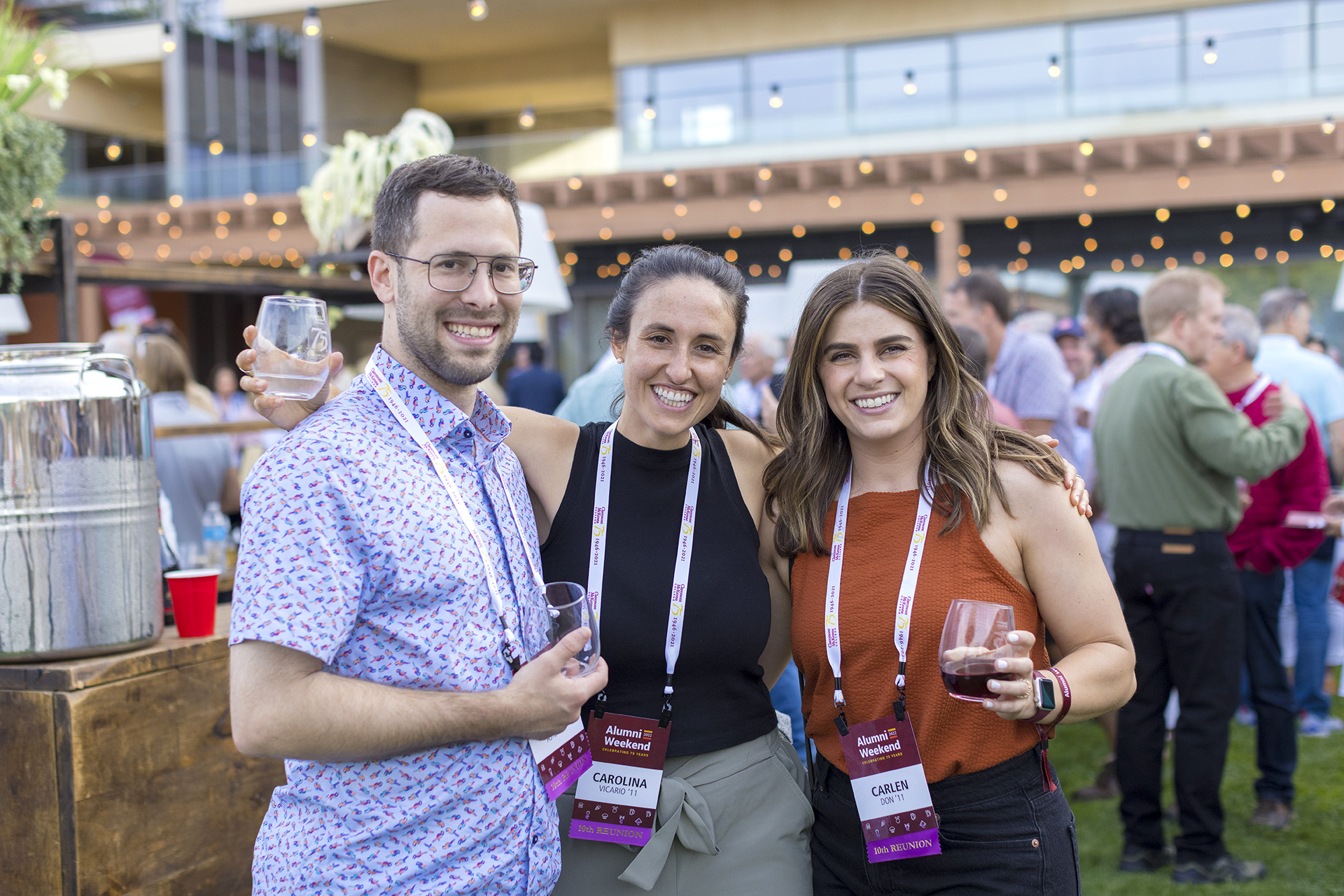 Three alumni pose together near the wine bar with their commemorative 75th Anniversary glasses during 2022 Alumni Weekend's wine tasting event.