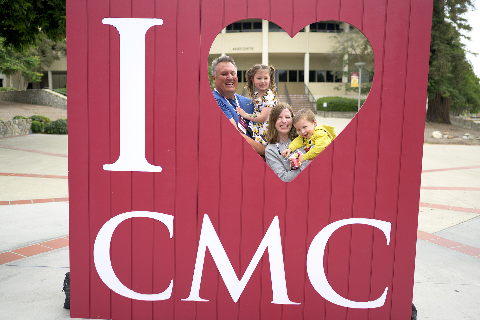 A family of alumni and their young children pose inside the heart-shaped cutout of the "I ♥︎ CMC" photo prop frame.