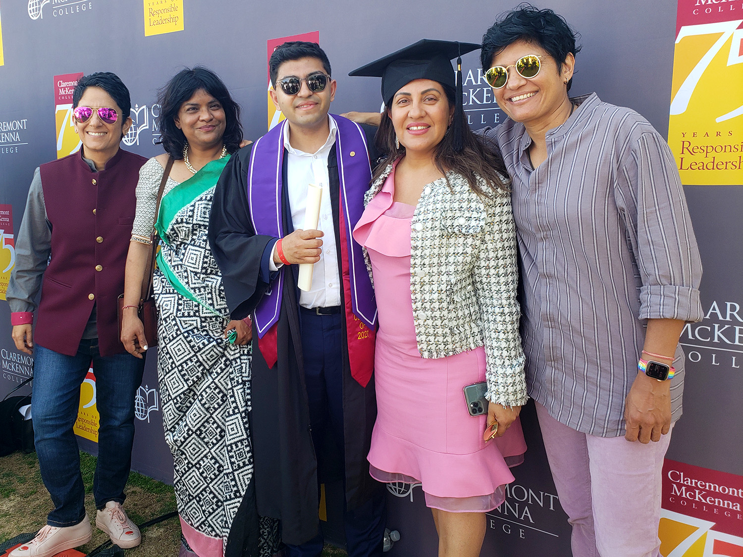 Sahib Bhasin ’21 and his family photographed together against the gray CMC 75th Anniversary logo pattern backdrop. His mother wears his mortar board hat.