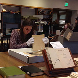 A CMC student works inside a historical archive, handling sheets of printed text with gloved hands.