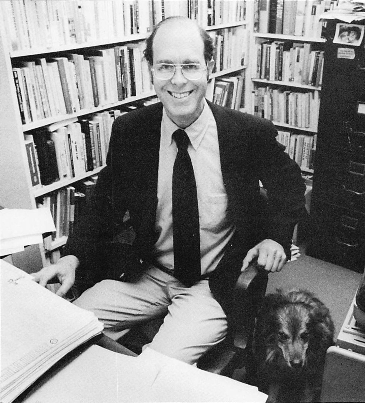 Black and white photo of Prof. Ward Elliott sitting in his office with his canine companion.