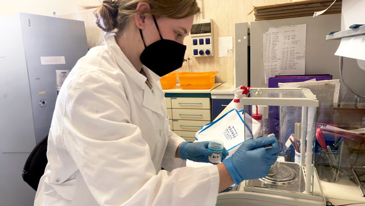 A student in a lab coat with a face mask in a laboratory working with a sample in a small jar
