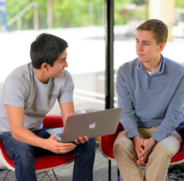 Two students sit talking, with a dark-haired male student holding a laptop and the other sits listening with his hands folded between his knees