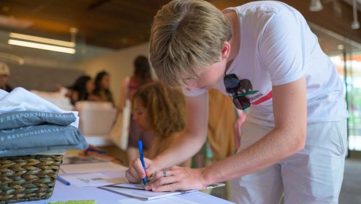 A student leans over a table, signing their name onto a sheet of paper