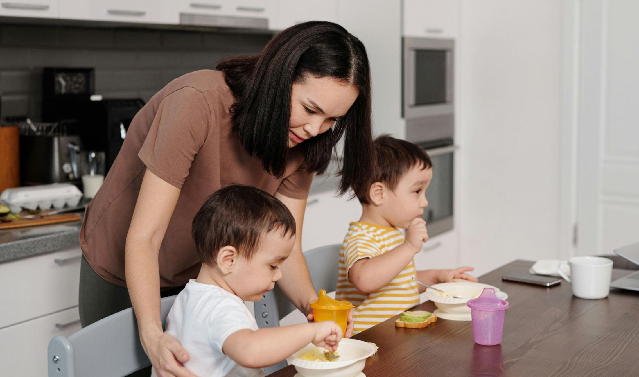 A mother standing behind her children eating at a table.
