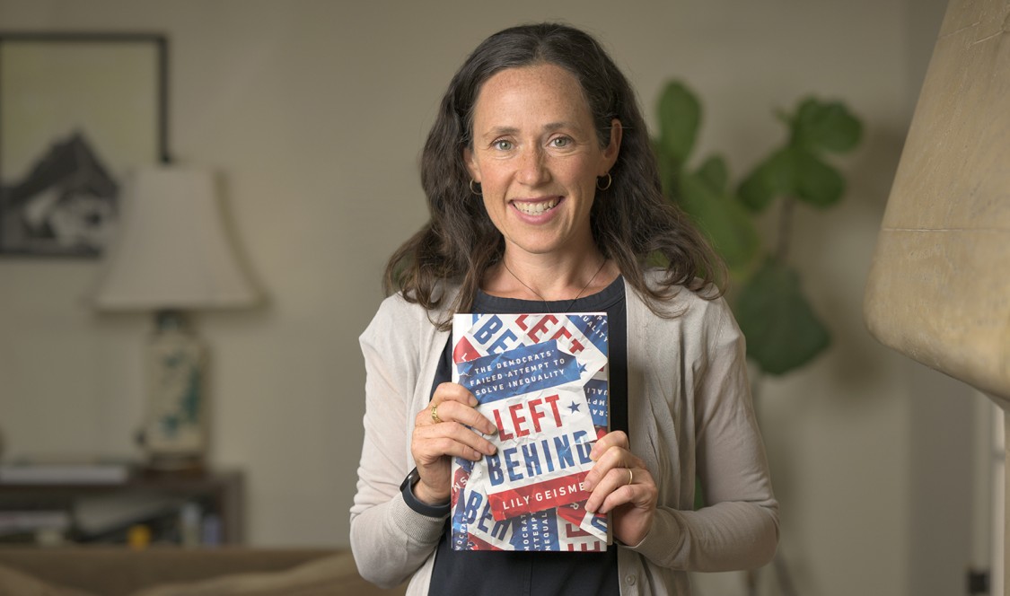 Professor Lily Geismer photographed at home, smiling and holding up her latest book, &quot;Left Behind.&quot;