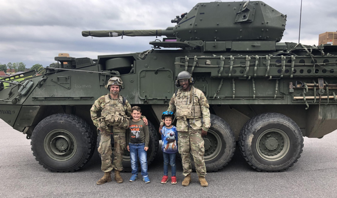 Ash and his friend stand with two children in front of a tank.