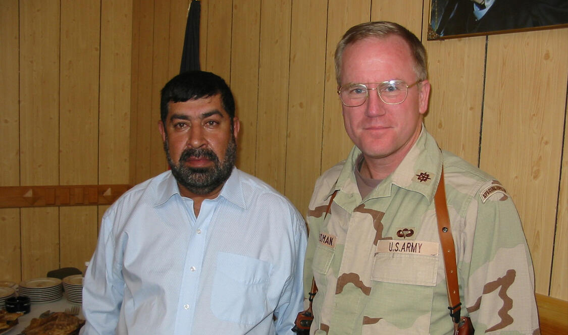 Two men standing in military uniform with glasses and white long sleeve top.