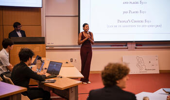 Woman in brown jumpsuit holding microphone speaking in front of students at a seminar.