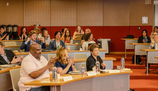 Group students and staff sitting at desks inside building clapping for speaker on stage.