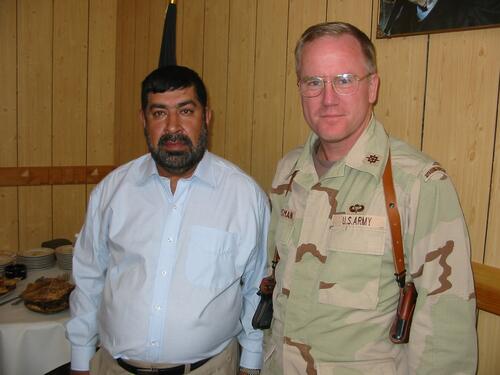 Two men, one with short black hair wearing white long sleeve button up and the other with short blonde hair wearing glasses and military uniform. 