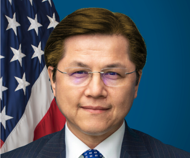 Headshot of Nury Turkel, a Uyghur man wearing glasses. He wears a suit with a blue patterned tie and stands in front of an American flag