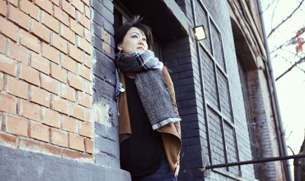 A photo of alumna Tracy Wang '04 leans against a building that's color coordinated with her cold weather outfit. A bare tree in the background indicates fall season.