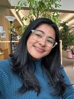 Woman in blue top wearing glasses with short black wavy hair sitting in front of books taking a selfie.